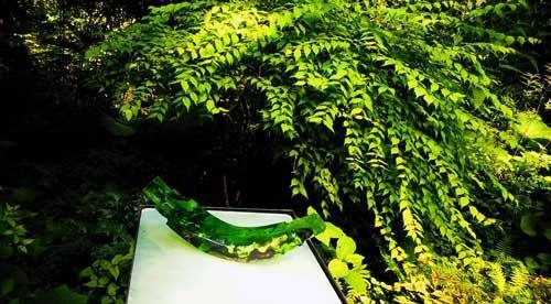 Pièce en pâte de verre verte posée devant un feuillage.